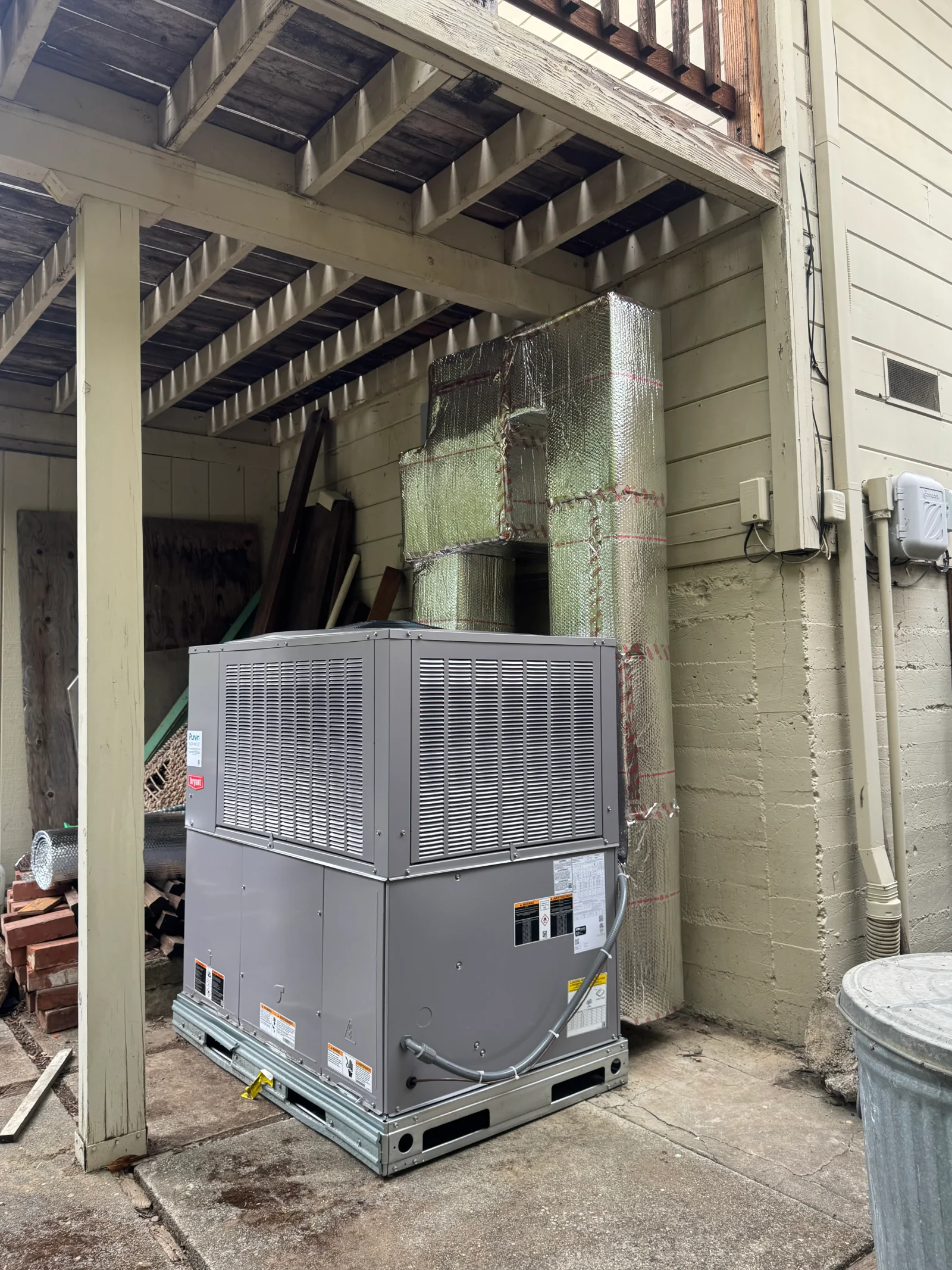 Gray packaged HVAC unit with louvered panels on concrete pad beside foil-wrapped insulated ductwork and exposed wood patio framing