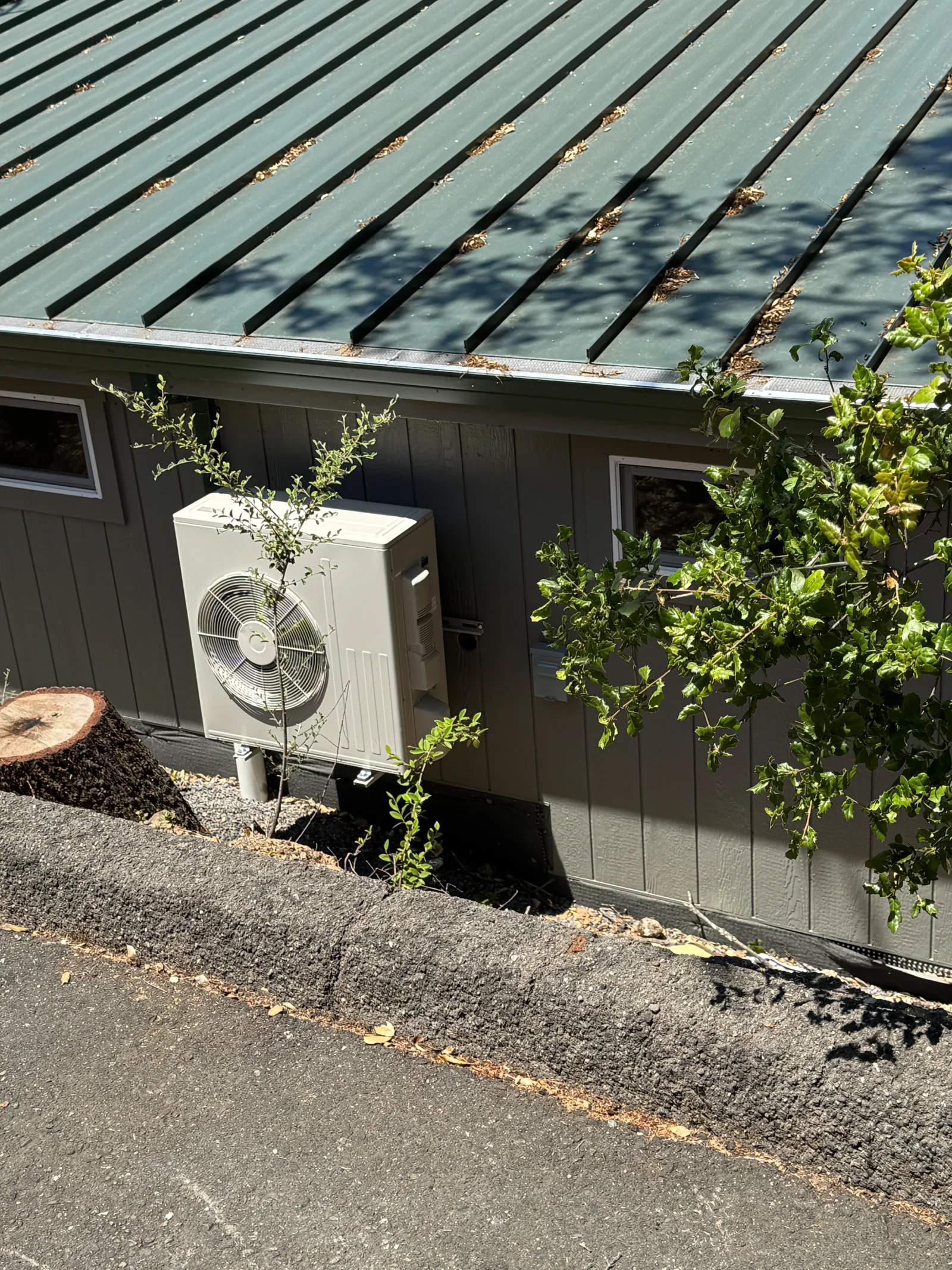 Wall-mounted ductless mini-split condenser with fan grille on gray board siding beside a green metal roof