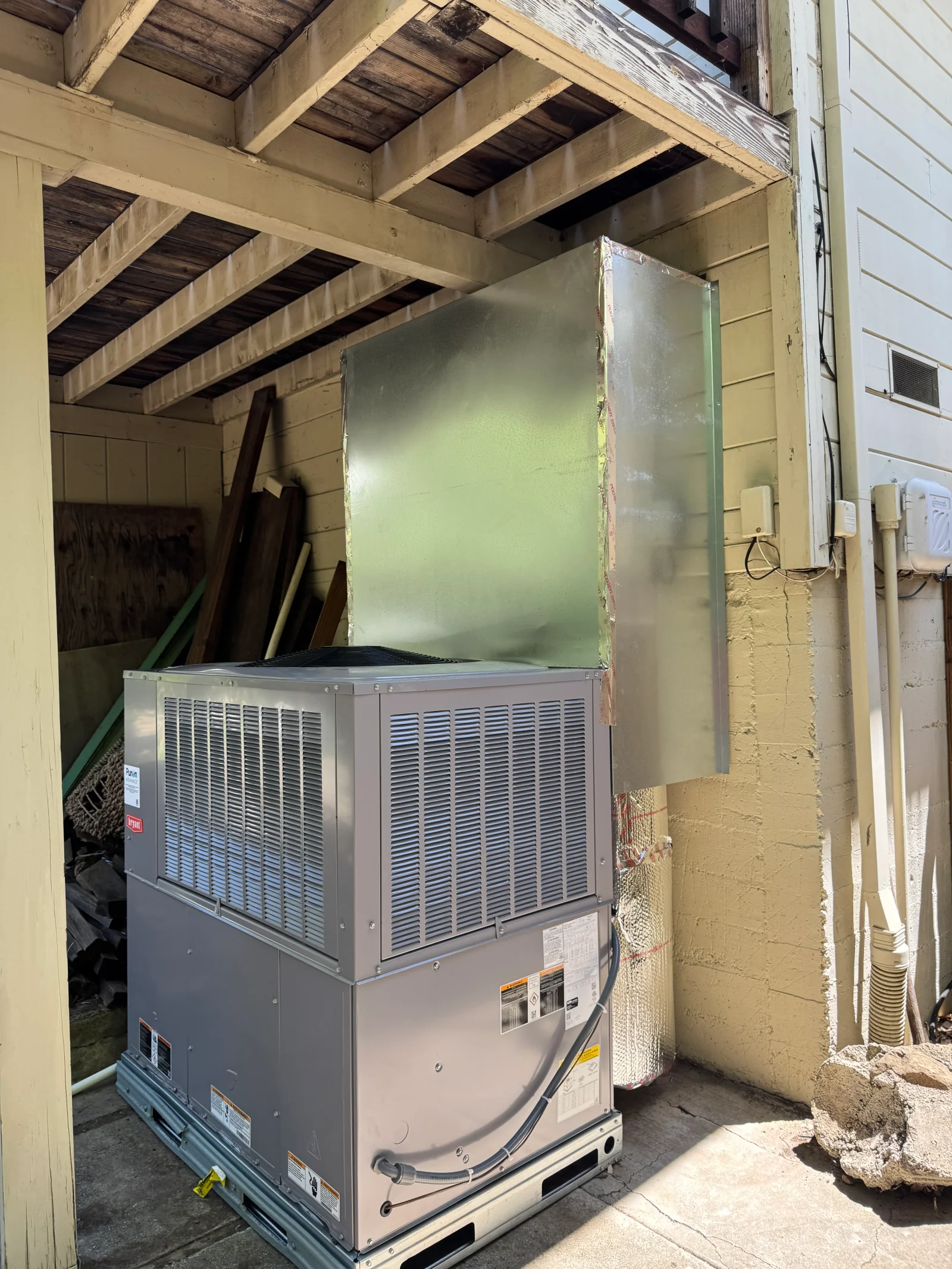 Gray Bryant packaged HVAC unit with louvered coils, galvanized sheet metal ductwork, and foil-wrapped insulated duct under a wood overhang